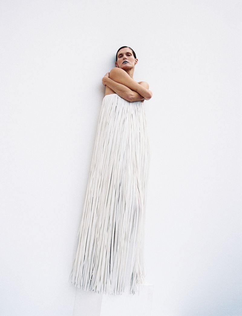 Fashionable adult female in a studio setting, wearing a long, white, fringed dress and makeup with a white wall as background.
