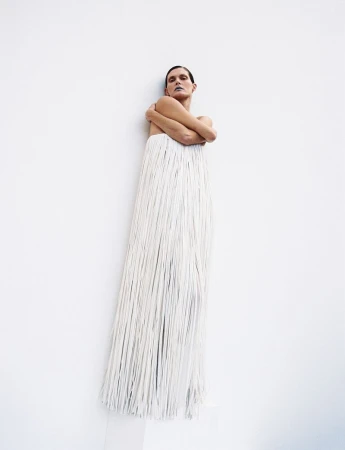 Fashionable adult female in a studio setting, wearing a long, white, fringed dress and makeup with a white wall as background.