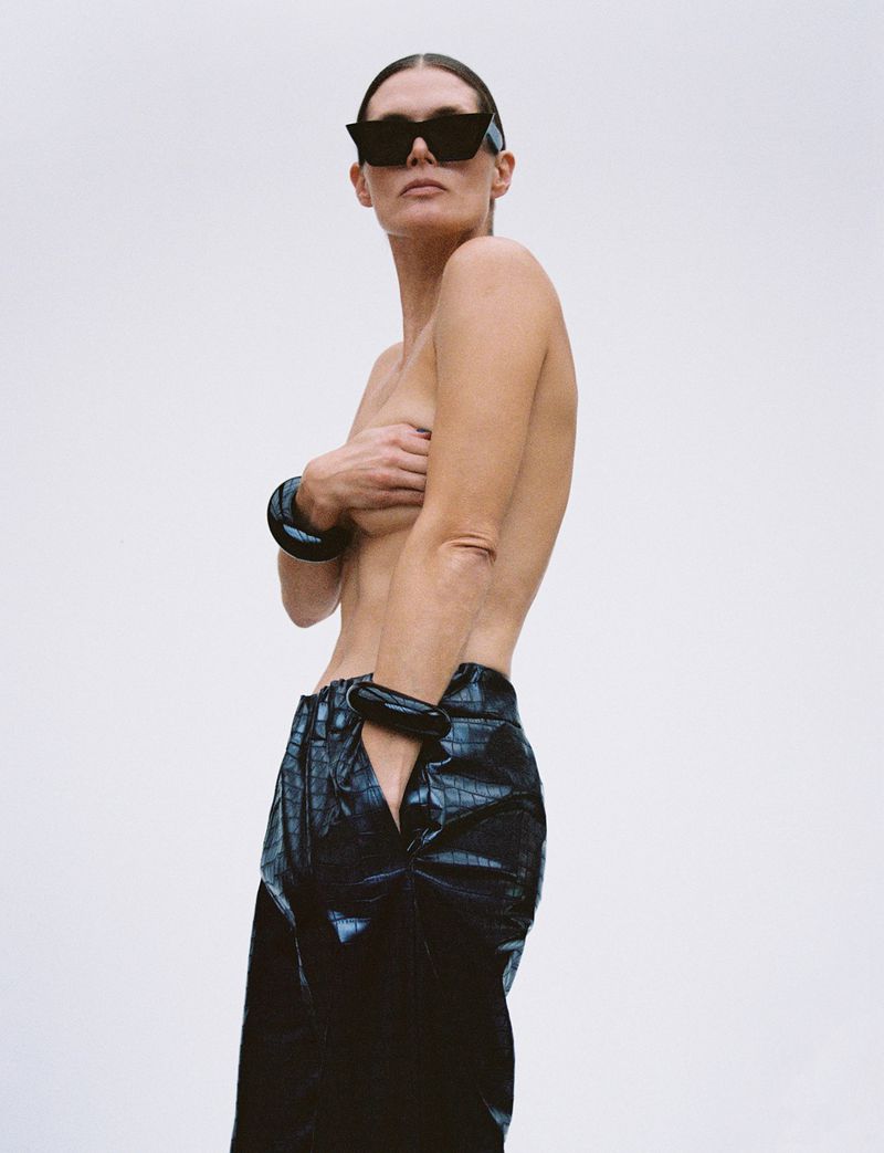 An adult female with bare chest poses in front of a plain white background, wearing sunglasses and bracelets and black reptile-pattern trousers.