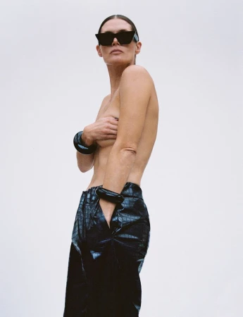 An adult female with bare chest poses in front of a plain white background, wearing sunglasses and bracelets and black reptile-pattern trousers.