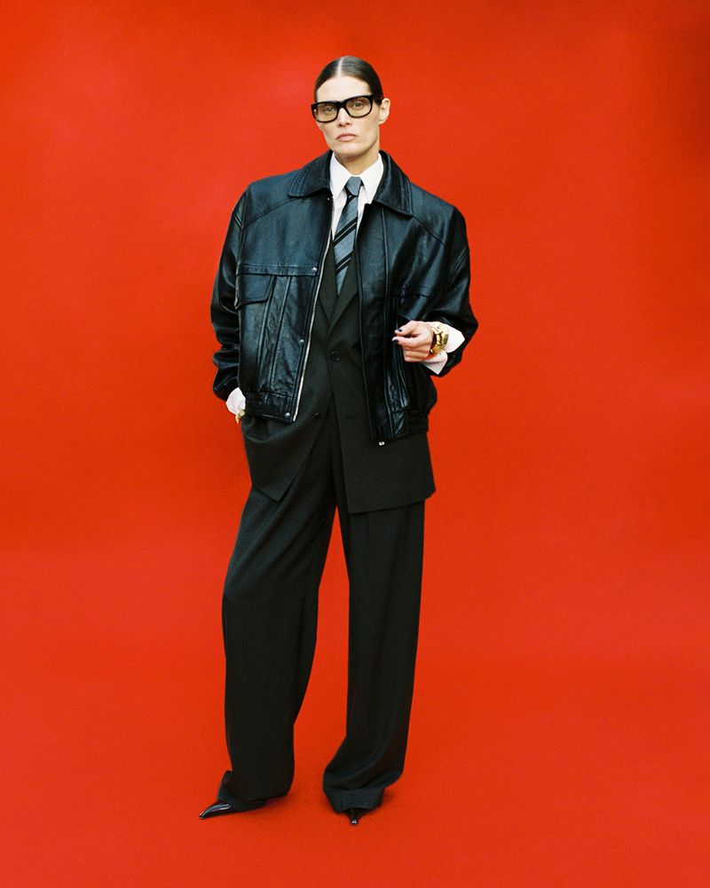 Woman in black suit and bomber jacket posing against red background, wearing glasses and rings, fashion-forward professional look.