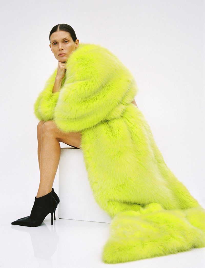 A woman sits in a studio wearing a large, fluorescent-yellow fur coat with black heeled booties against a clean, white backdrop.