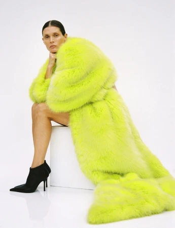 A woman sits in a studio wearing a large, fluorescent-yellow fur coat with black heeled booties against a clean, white backdrop.