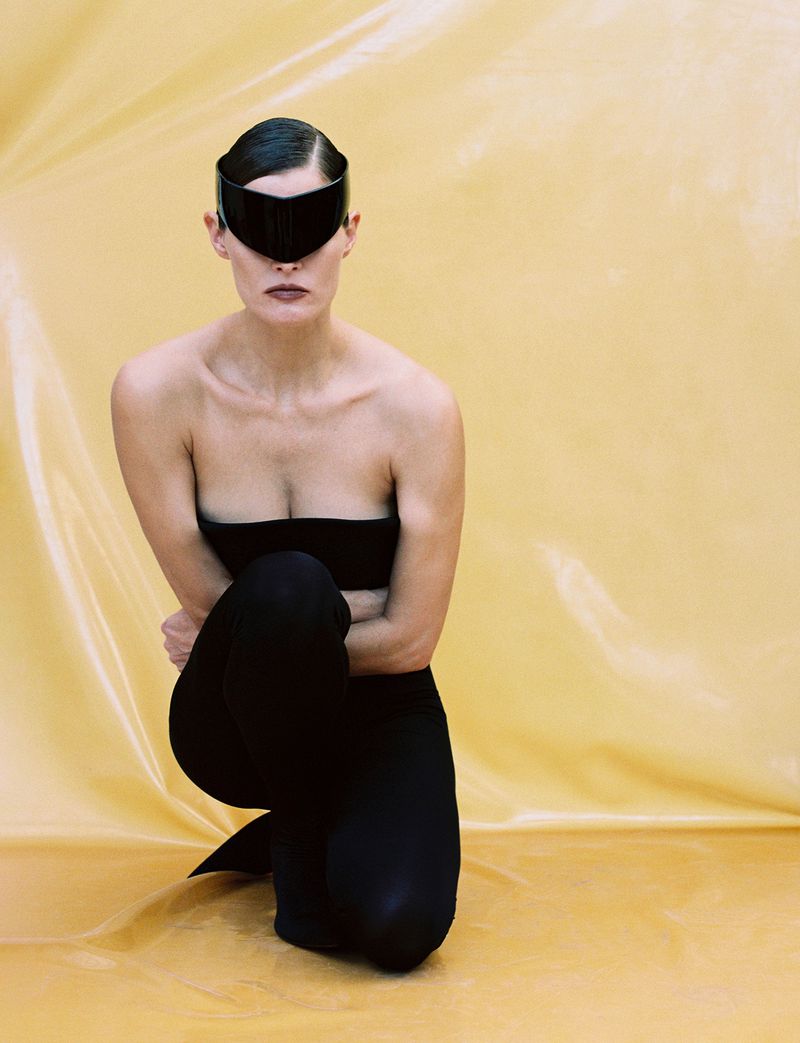 A woman is kneeling in a studio setting, wearing black clothing and a futuristic black visor against a yellow-toned backdrop.