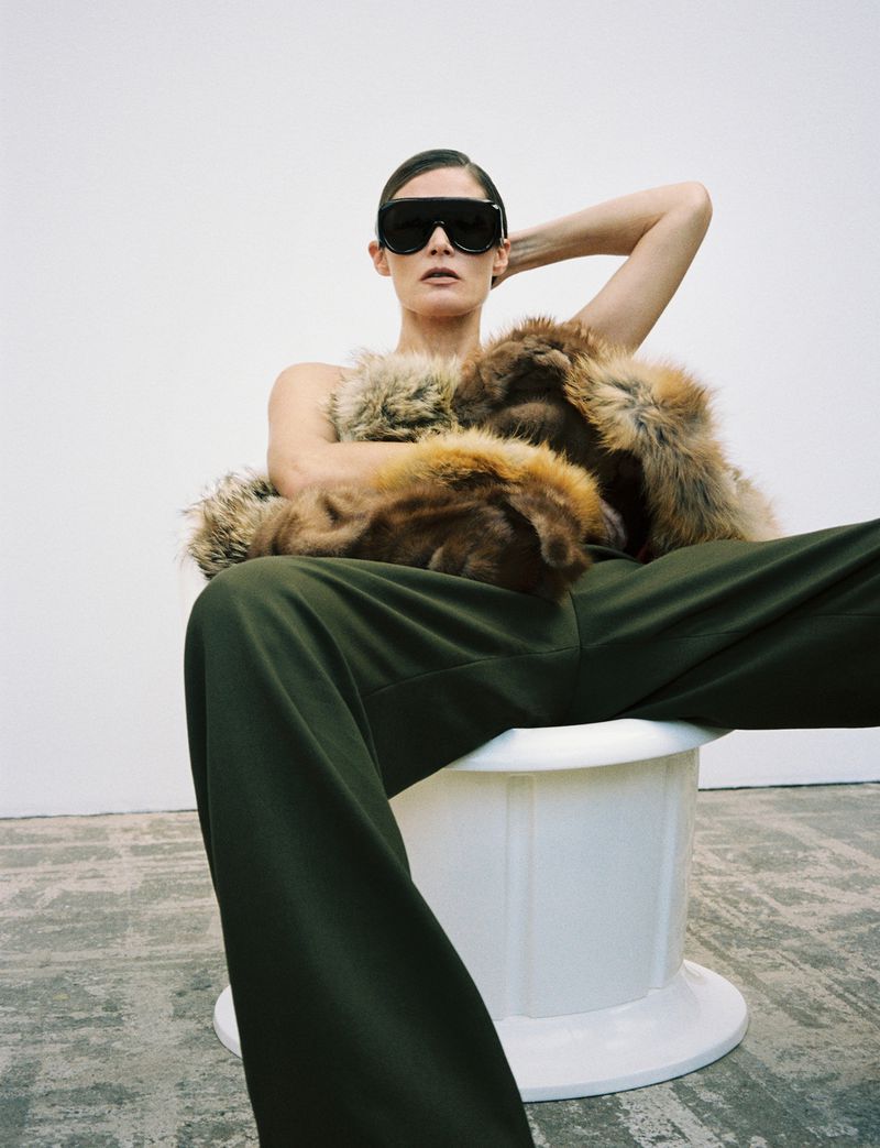 Elegant woman in sunglasses and a fur stole posing indoors. She wears dark green pants and sits on a white pedestal.