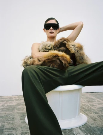 Elegant woman in sunglasses and a fur stole posing indoors. She wears dark green pants and sits on a white pedestal.