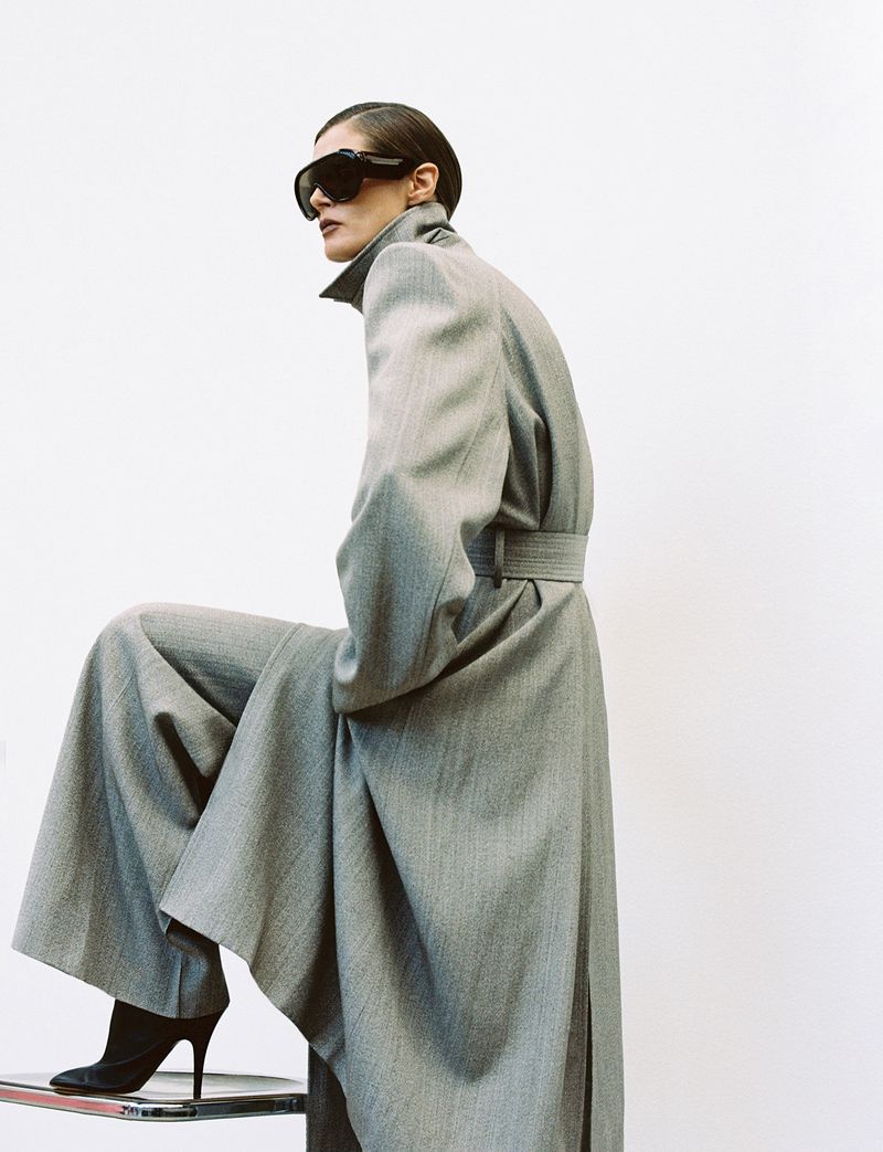 A fashion-forward woman poses in a light gray belted coat and wide-leg pants, wearing dark sunglasses, set against a plain white backdrop.