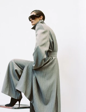 Model Malgosia Bela wearing a grey coat and oversized sunglasses, posing with one foot on a stool against a white background.