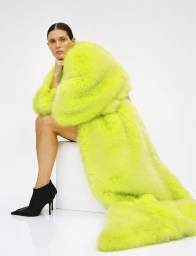 Model Malgosia Bela seated on a white block wearing a voluminous neon yellow coat and black boots against a white backdrop.