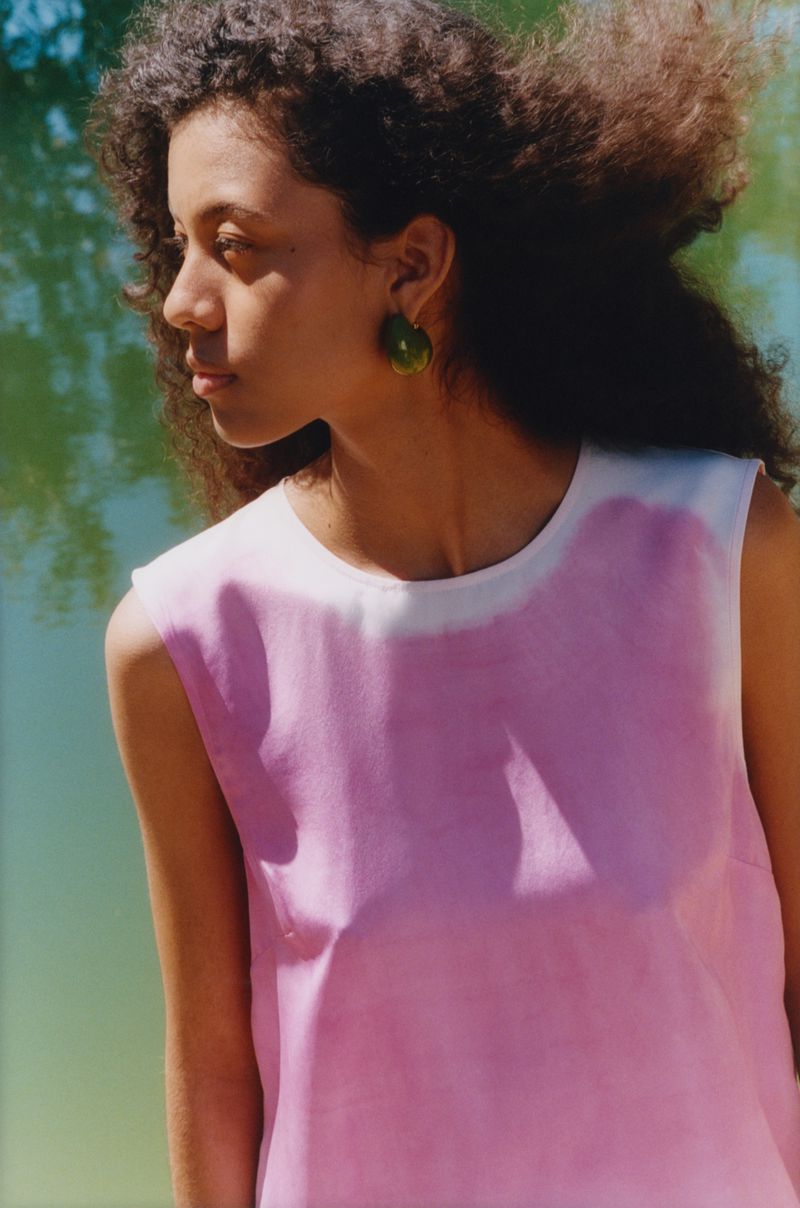 Young woman in a sleeveless top gazes left near a body of water, bathed in sunlight, showcasing an elegant green earring and natural curly hair.