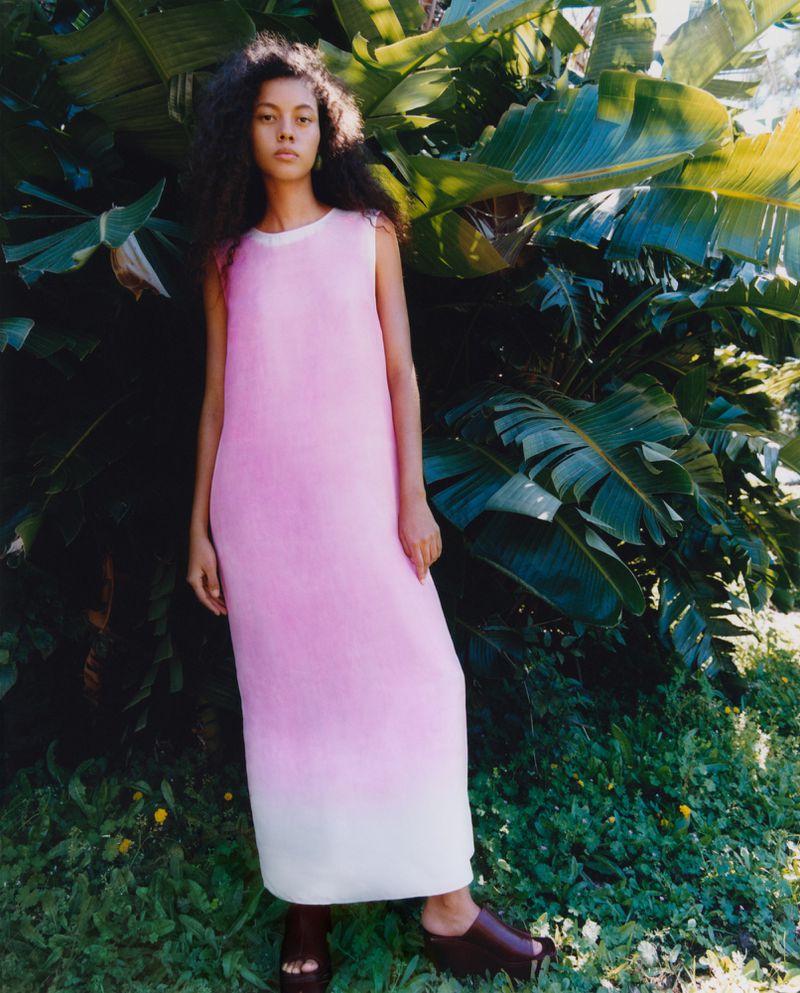A woman with curly dark hair stands outdoors in a pink and white sleeveless dress, posing in front of lush banana plants.