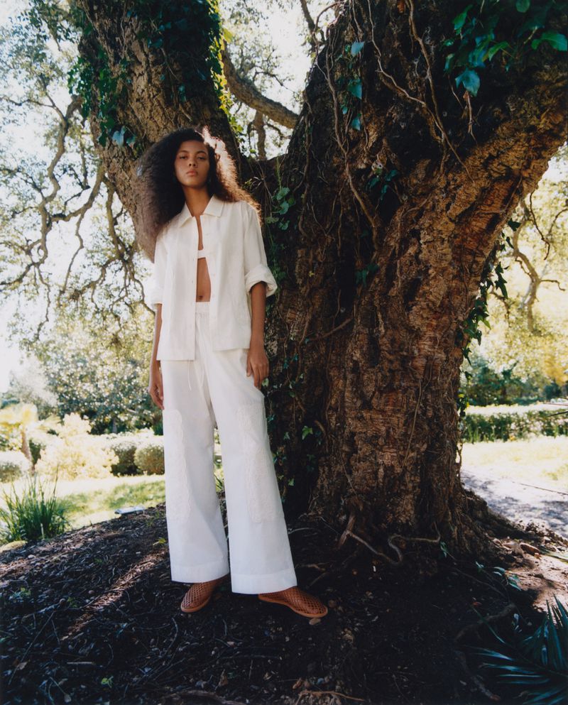 Woman in white linen outfit poses against large tree in outdoor setting, capturing a blend of fashion and natural backdrop.