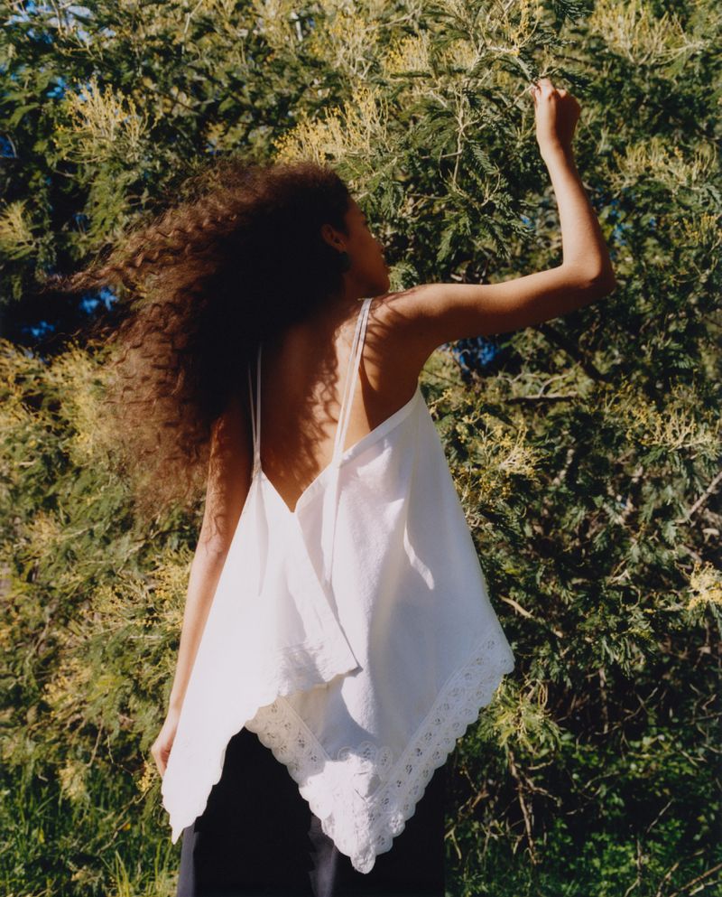 Woman with curly hair in white top outdoors reaching to a tree. Back and arm are visible. Soft natural light. Fashion photography.