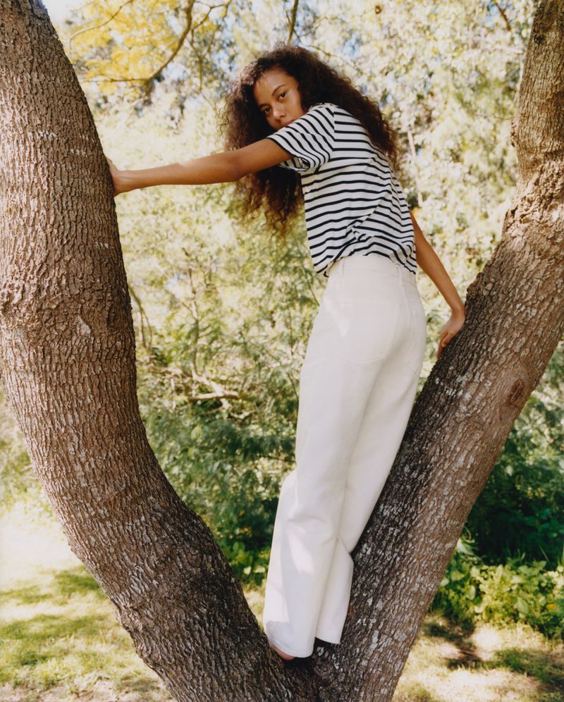 Adult woman with dark curly hair climbs in a tree wearing a striped t-shirt and white pants in an outdoor environment with green foliage.