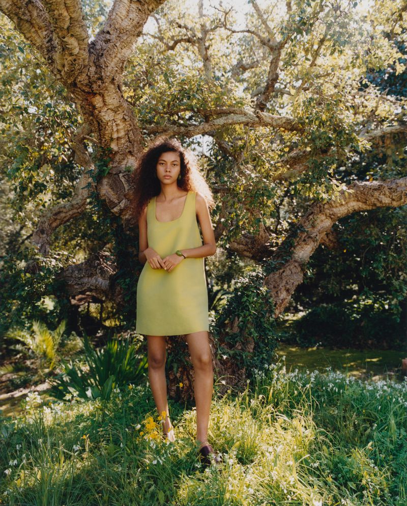 A young woman in a chartreuse dress stands in a lush green meadow surrounded by trees, illuminated by soft, natural light.