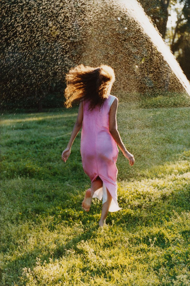 Back view of woman in pink dress running through grass under sprinkler. Wet curly hair. Sunlight and water drops in outdoor setting.