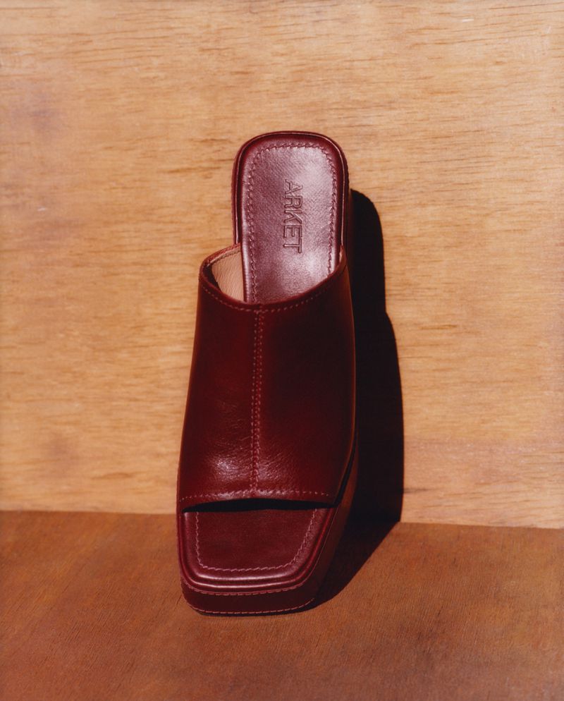 Studio shot of a burgundy leather heeled mule by ARKET on brown and light wood backdrop, showcasing fashion footwear in a minimalist style.