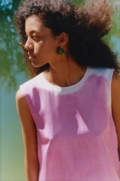 Model with dark curly hair wearing a pink sleeveless top against an outdoor water background for Arket SS25.