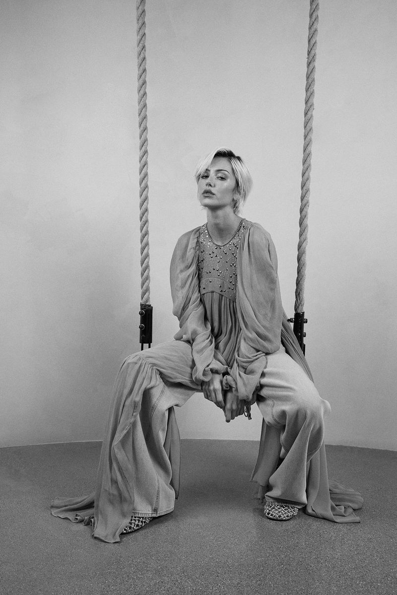 Black and white portrait of a woman in a dress and pants, sitting on a rope swing against a neutral wall, looking at the camera.