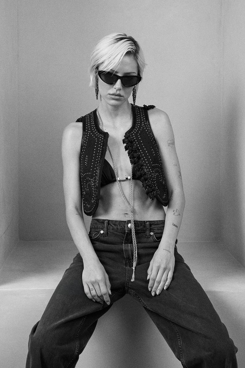 A young adult woman with blonde hair, wearing a black bikini top, vest, and jeans, is sitting indoors against a plain background.