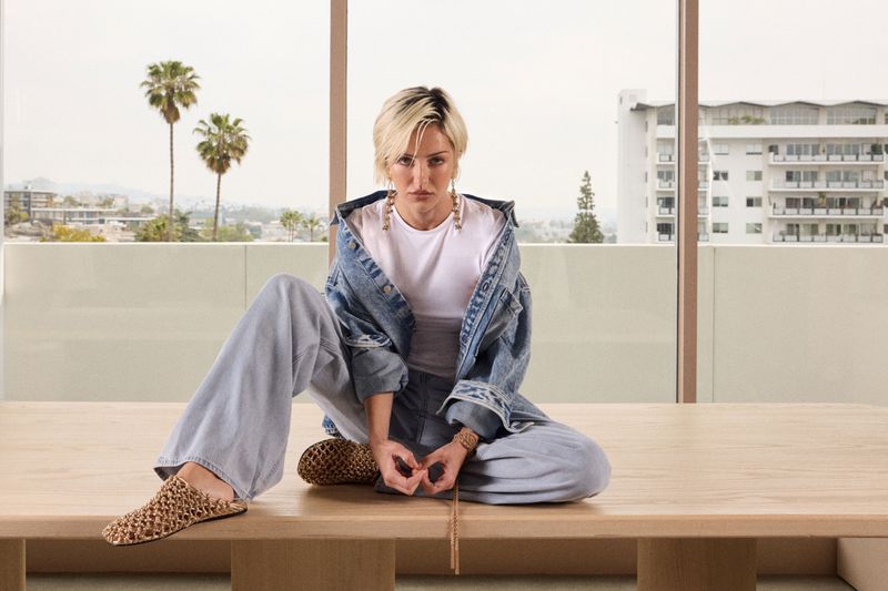 Adult woman in denim jacket and light blue jeans sits cross-legged on a table in front of a bright window with city view and palm trees.