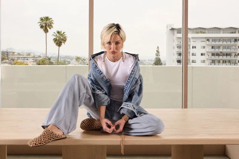 Adult woman in denim jacket and light blue jeans sits cross-legged on a table in front of a bright window with city view and palm trees.