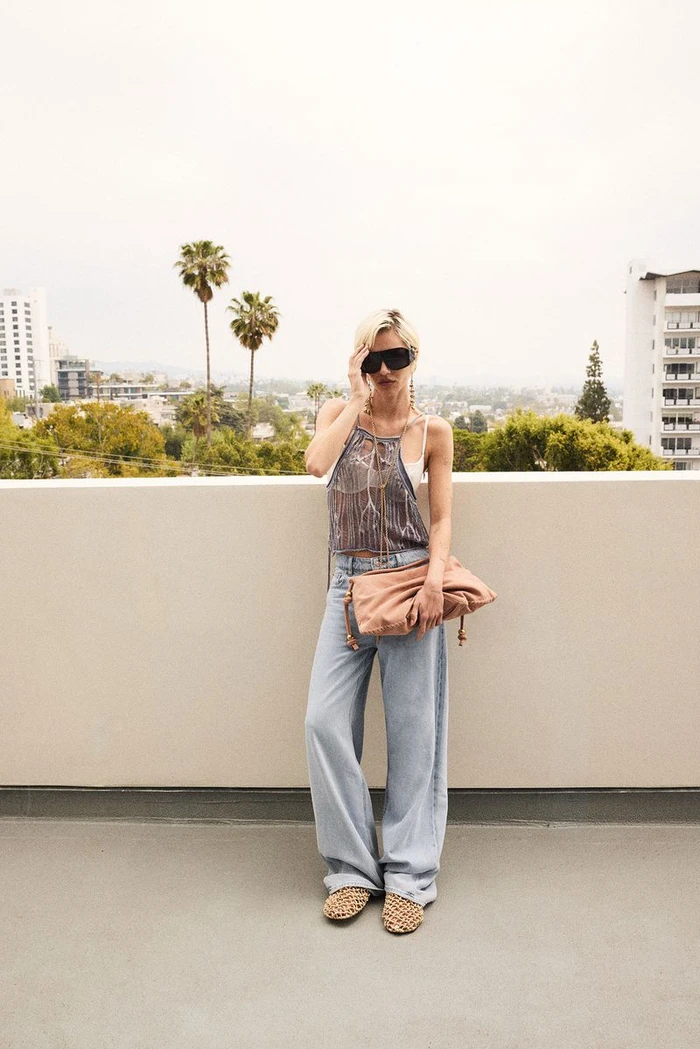 Model in oversized sunglasses wearing a patterned halter top, light wash jeans, and holding a pink clutch on a rooftop.
