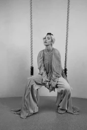 Model seated on a rope swing wearing long flowing trousers and a detailed top against a studio background.