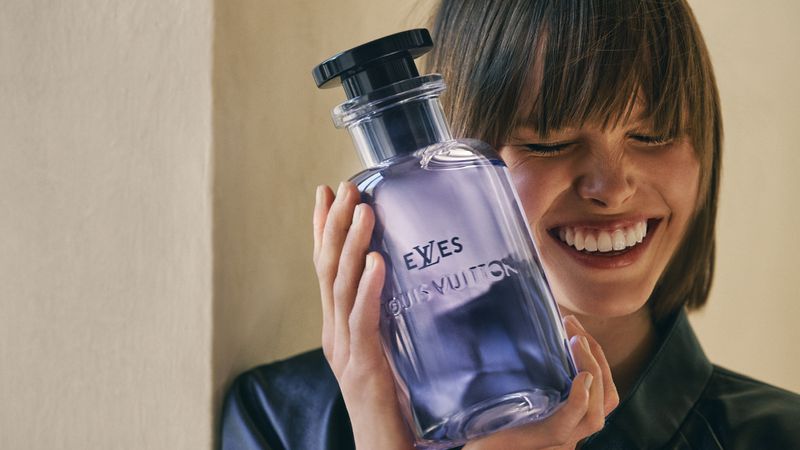 Smiling adult woman holds a translucent purple Louis Vuitton perfume bottle with a black cap in an indoor setting with soft lighting.