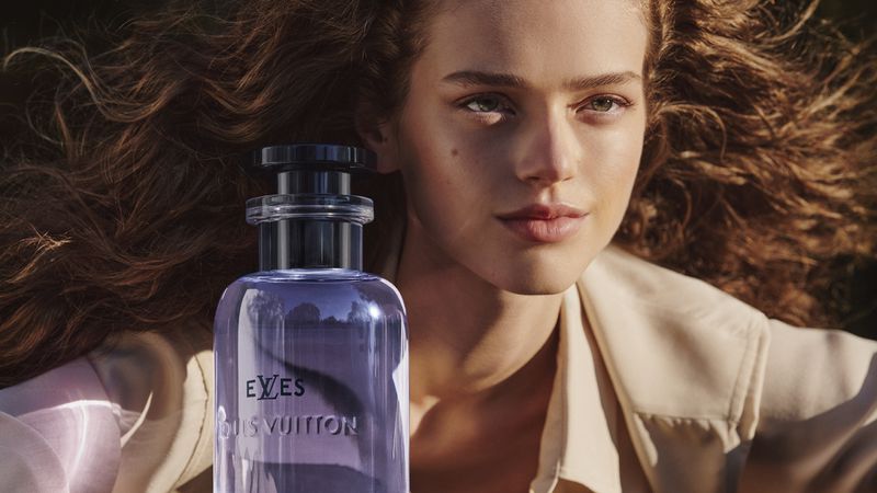 A woman with wavy auburn hair poses beside a Louis Vuitton perfume bottle in a close-up shot highlighting beauty and luxury.
