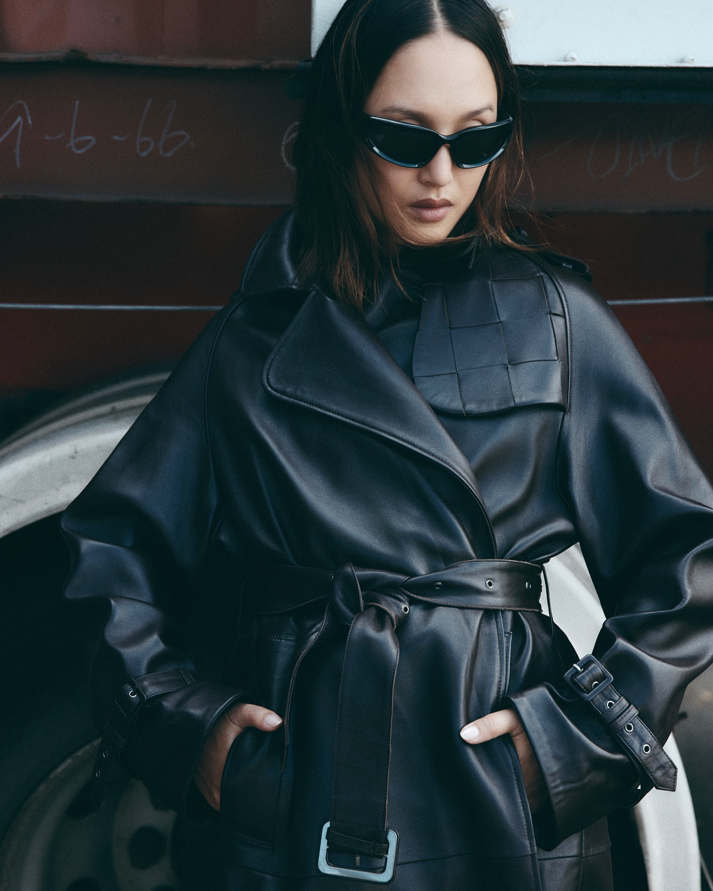 A woman in a black leather trench coat and sunglasses poses outdoors against a metal backdrop with graffiti-like markings and a tire visible.