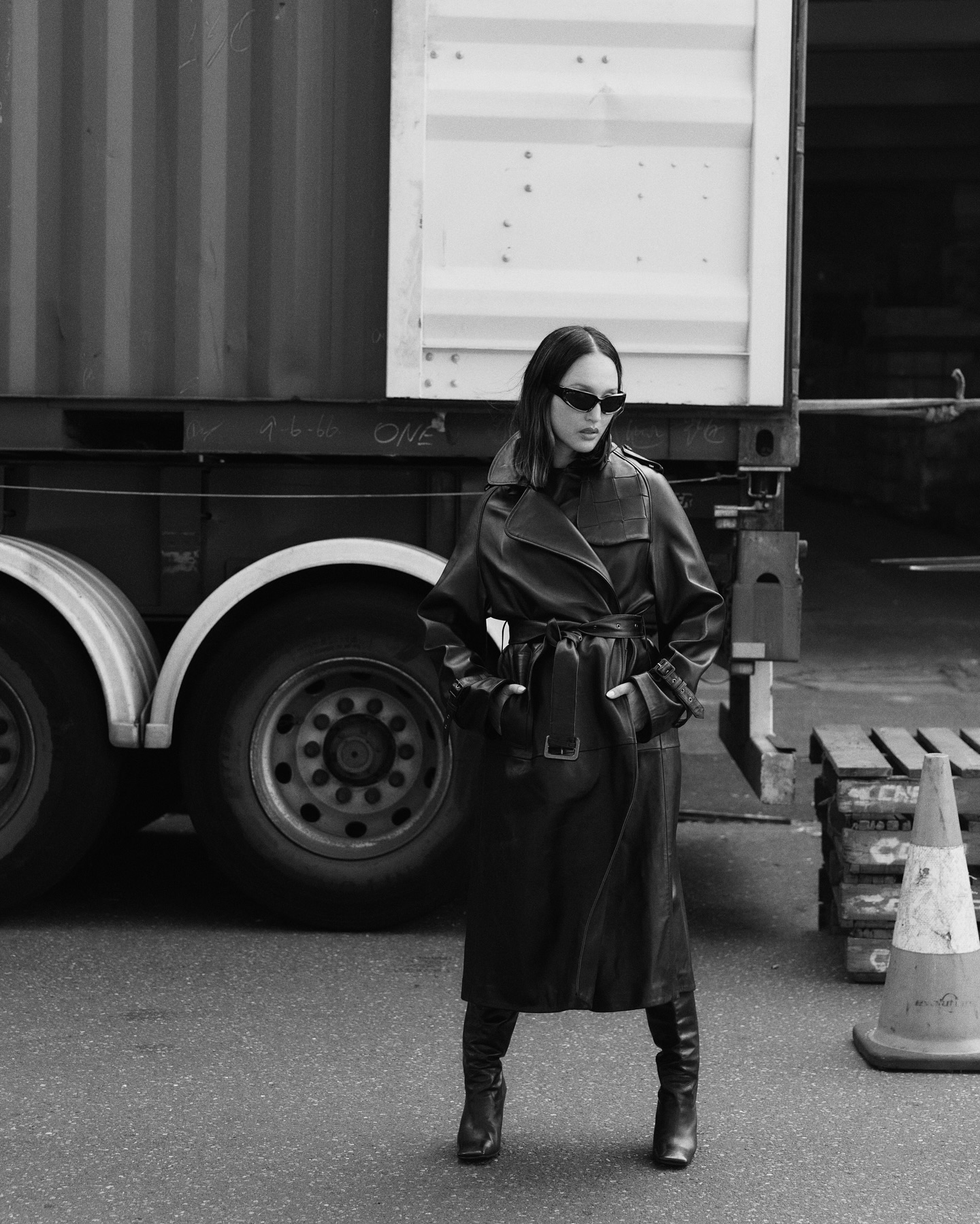 A woman in a trench coat and boots poses outdoors in a monochrome fashion shot with a truck and traffic cones in the background.