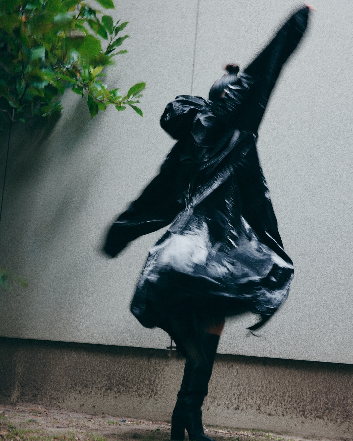 Motion blurred figure in a black coat and boots posing against a plain wall with foliage in the corner