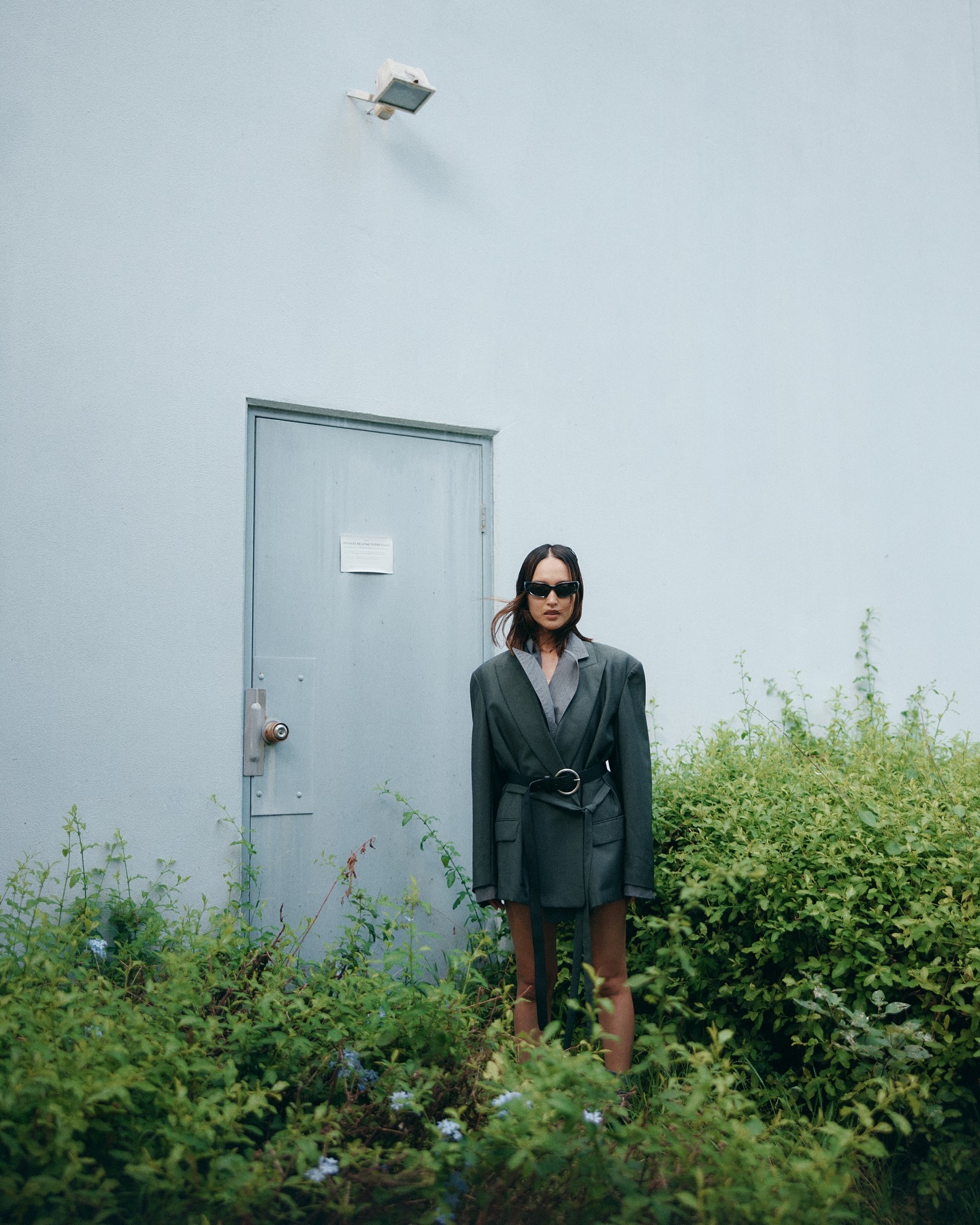 Adult female wearing blazer and sunglasses stands amongst plants in front of light-blue door and wall outdoors, eye-level angle.