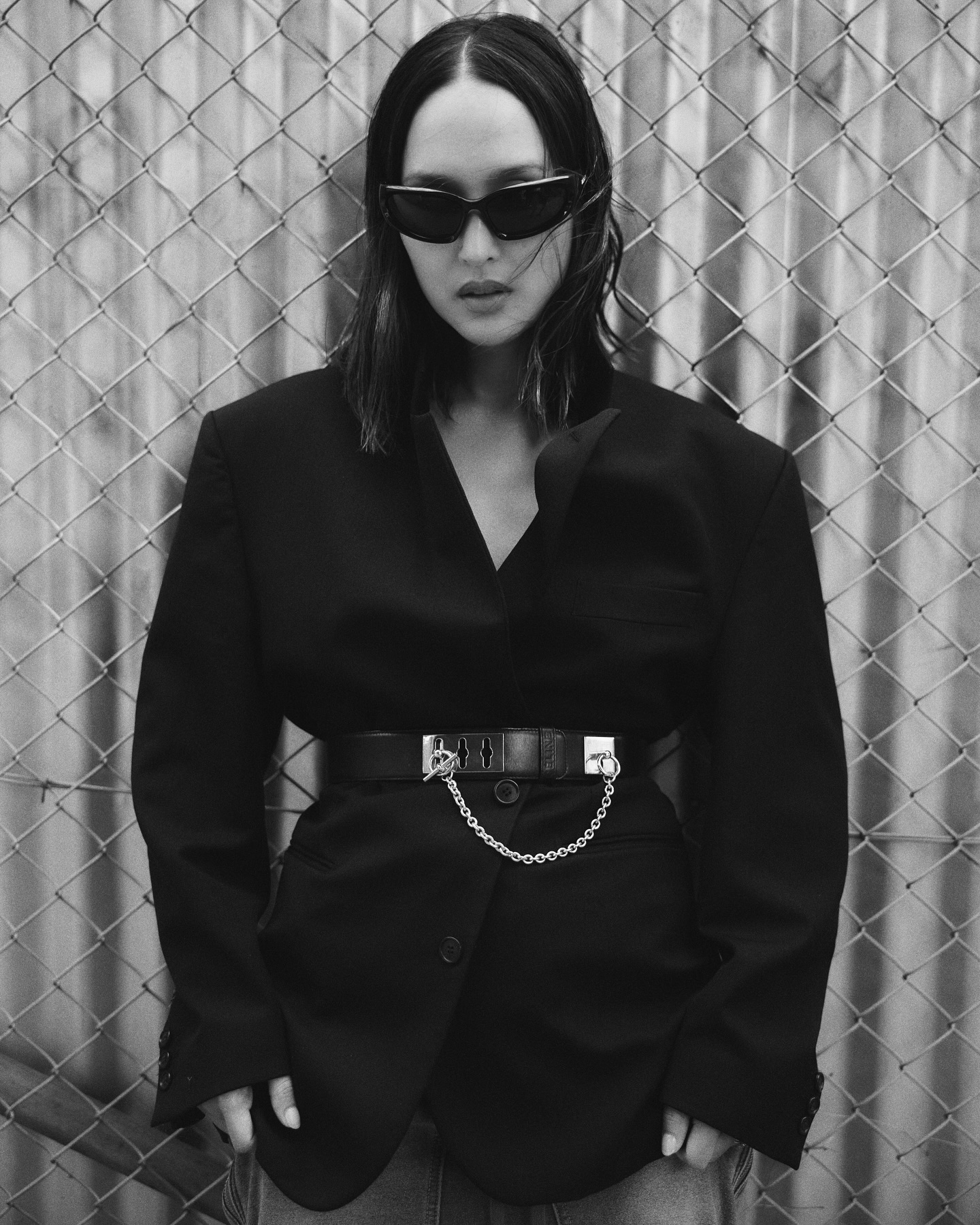 Black and white fashion portrait of a woman wearing sunglasses, a black blazer and jeans, posed against a chain-link fence.