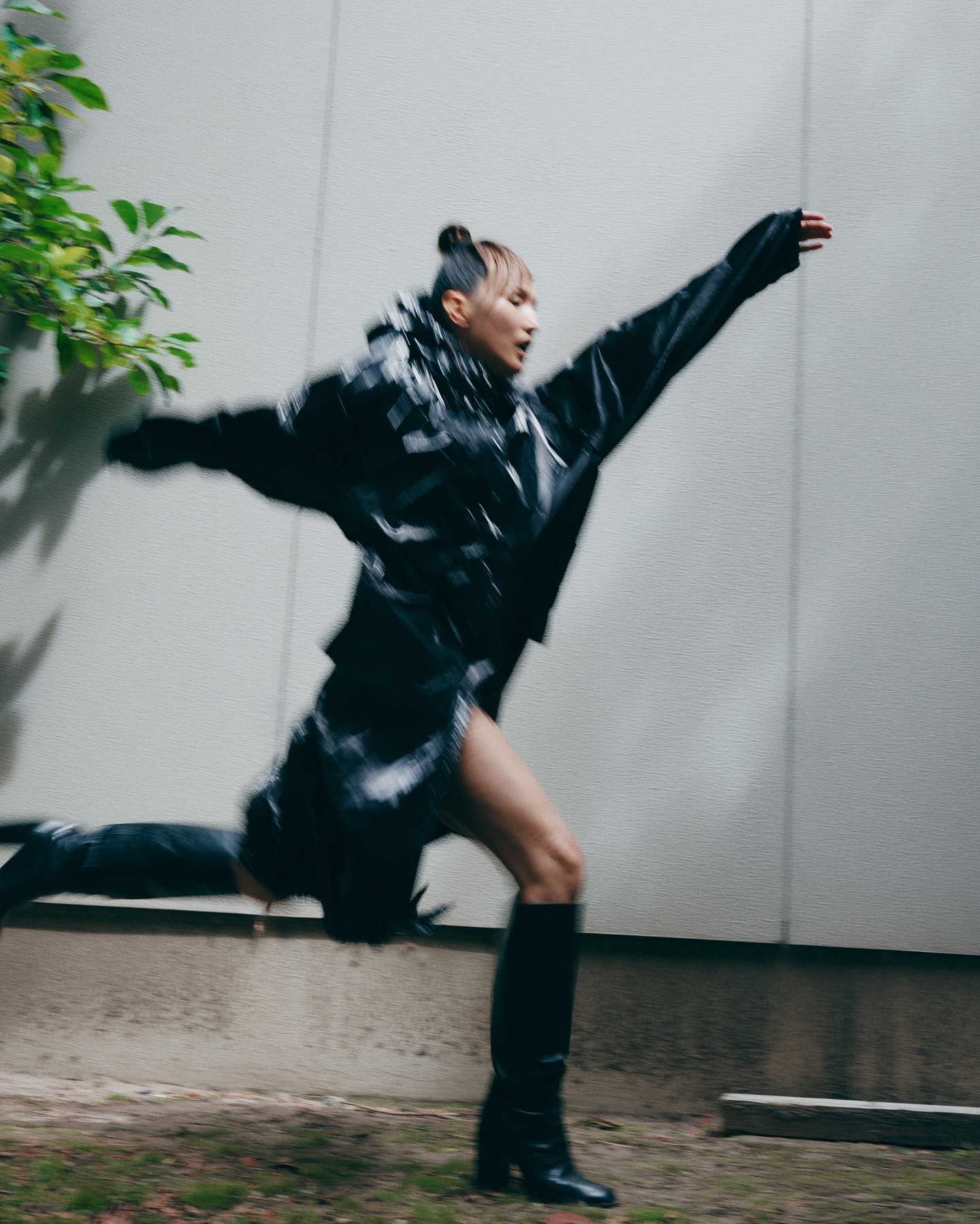 A woman in a black jacket and tall boots leaping across a light concrete exterior wall in motion blur.