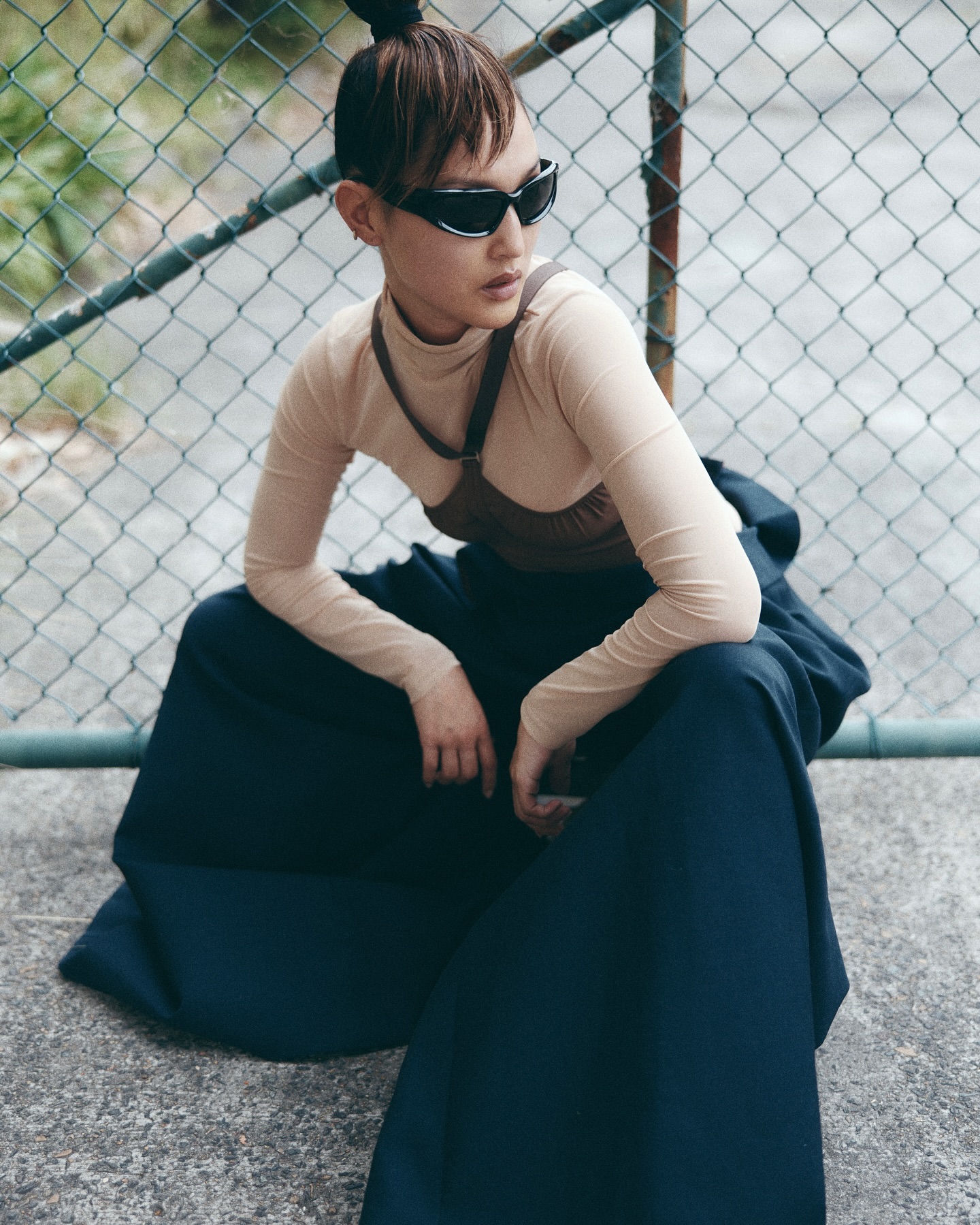 A stylish Asian woman with sunglasses sits in a beige top and blue skirt, framed by a chain-link fence and asphalt ground.