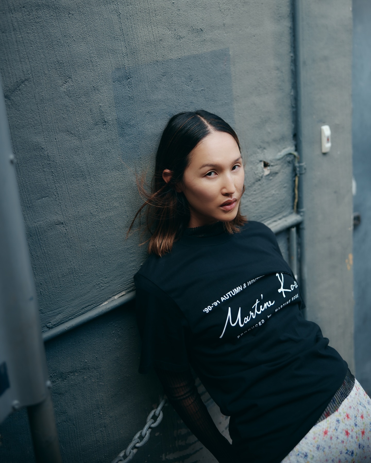 Woman leans against a gray wall wearing a black graphic tee and a floral patterned skirt in a natural outdoor setting.