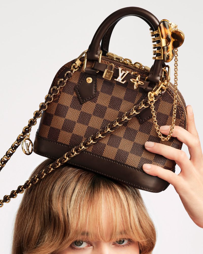 Brown checkerboard handbag by Louis Vuitton on top of a fair-skinned female’s head with bangs, with gold hardware details.