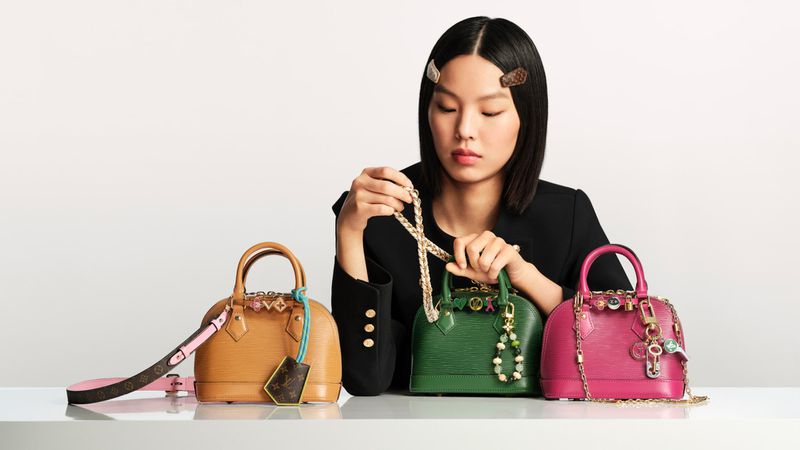 A woman presents an array of colorful Louis Vuitton Alma handbags, accessorized with charms and chain straps, against a bright studio backdrop.