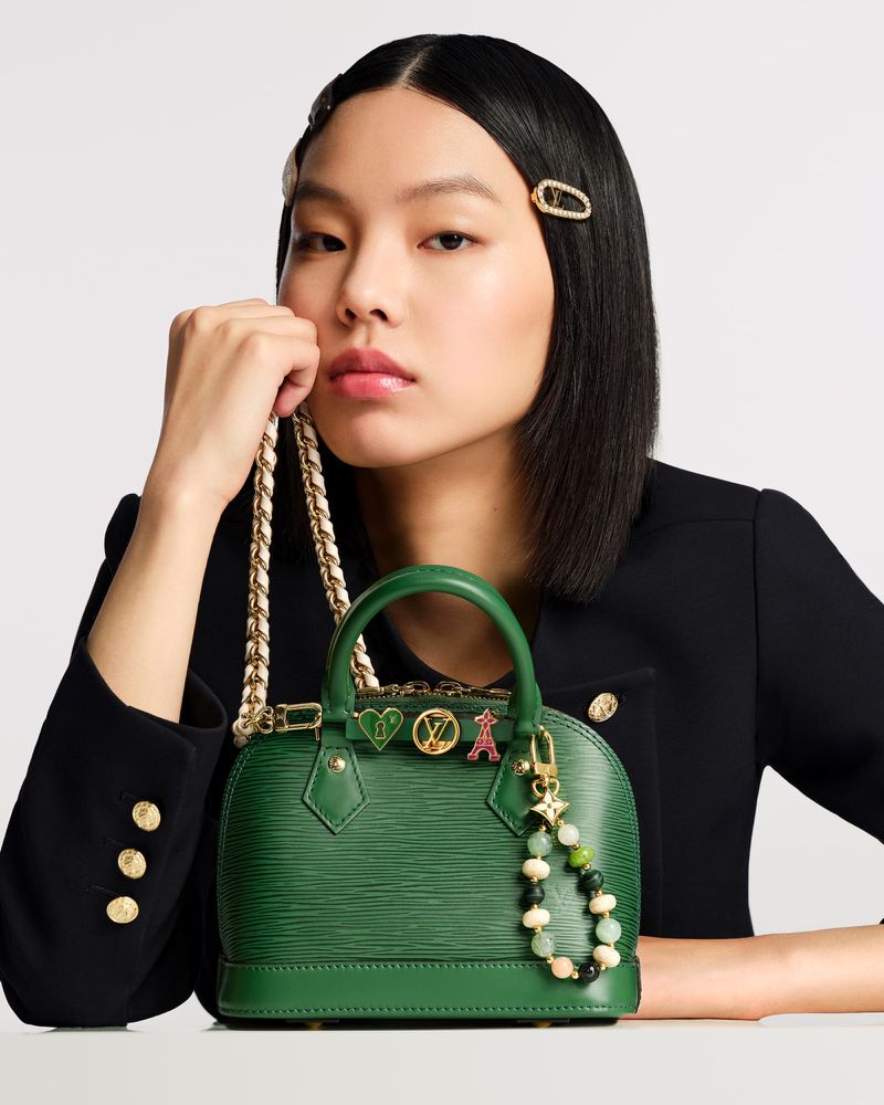Elegant Asian woman presents a small green Louis Vuitton purse with gold chain strap and decorative charms in a fashion studio shot.