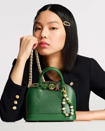Elegant Asian woman presents a small green Louis Vuitton purse with gold chain strap and decorative charms in a fashion studio shot.