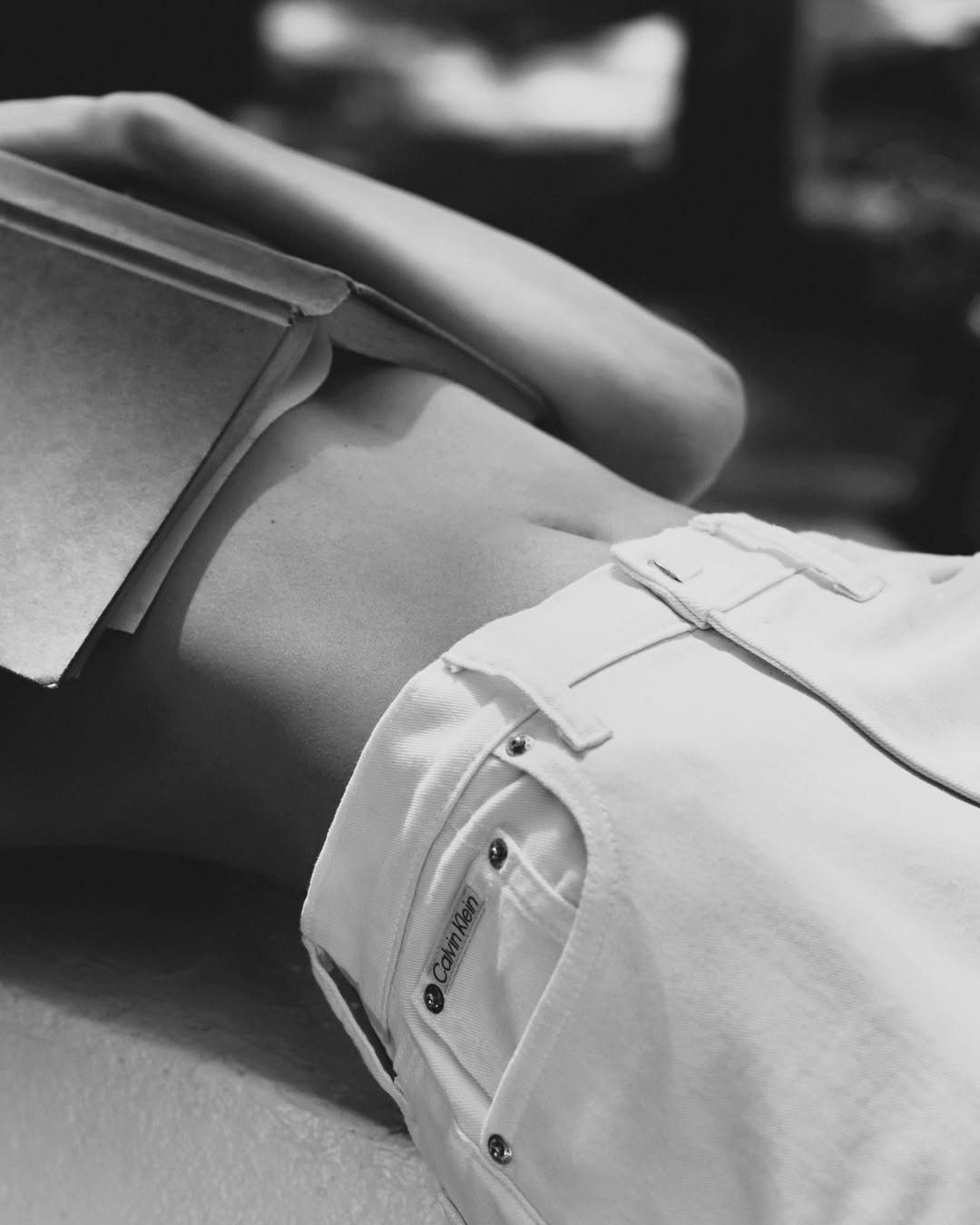 Black and white close-up of a model reclining with an open book resting on their torso, wearing white Calvin Klein denim jeans.