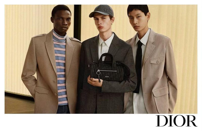 Three male models wearing stylish blazers and holding a Dior handbag in a studio with a yellow backdrop for a fashion editorial.
