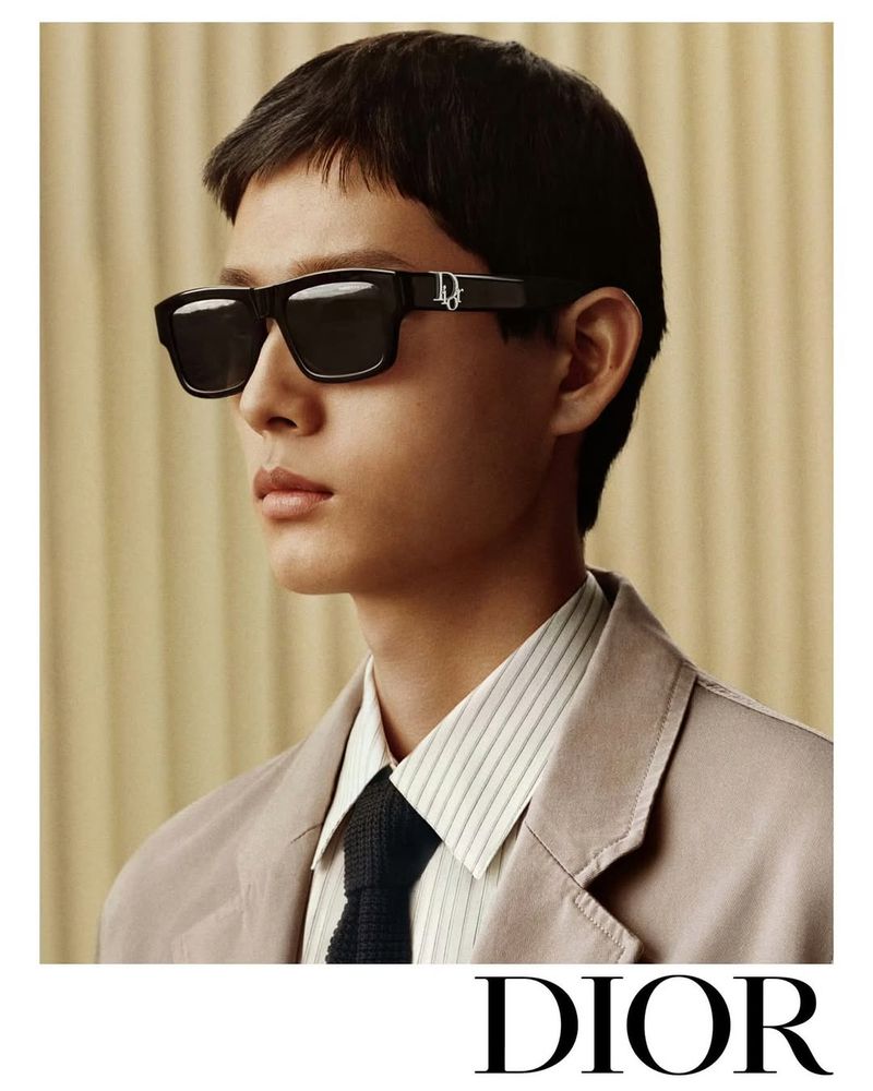 Portrait of a dark-haired male model wearing black sunglasses, a striped shirt, and a tan jacket with the Dior logo visible.