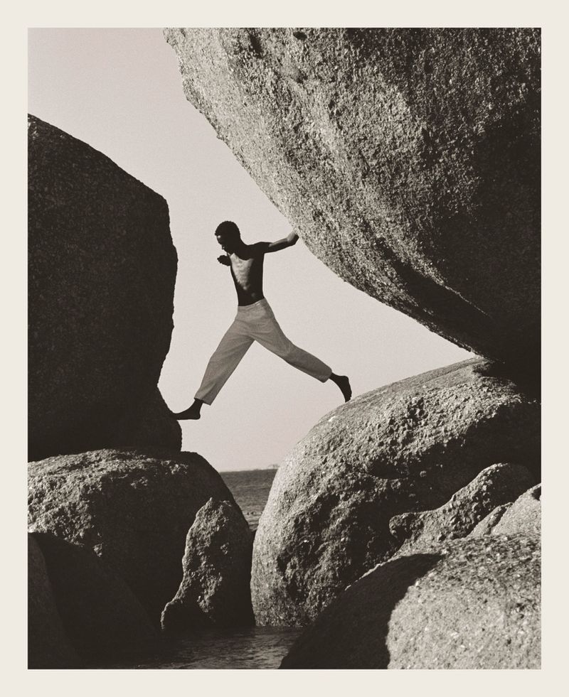 Man leaps between rock formations near water. The image is a black and white outdoor shot with neutral daylight.