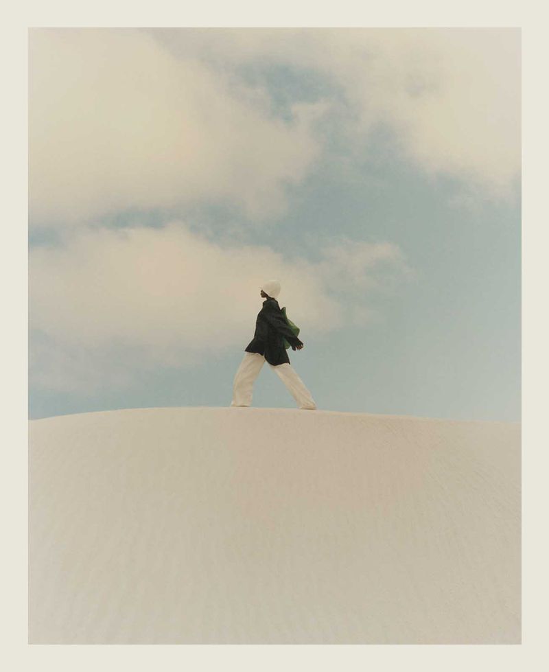 A person in a coat and pants walks on a dune against a blue and cloudy sky. The person's posture is relaxed and casual.