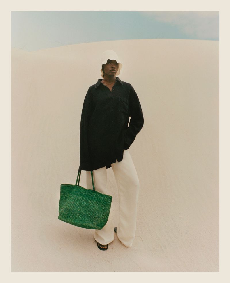 A woman stands on a sandy beach wearing a black top, white pants, and a white hat, and she is holding a green tote bag.