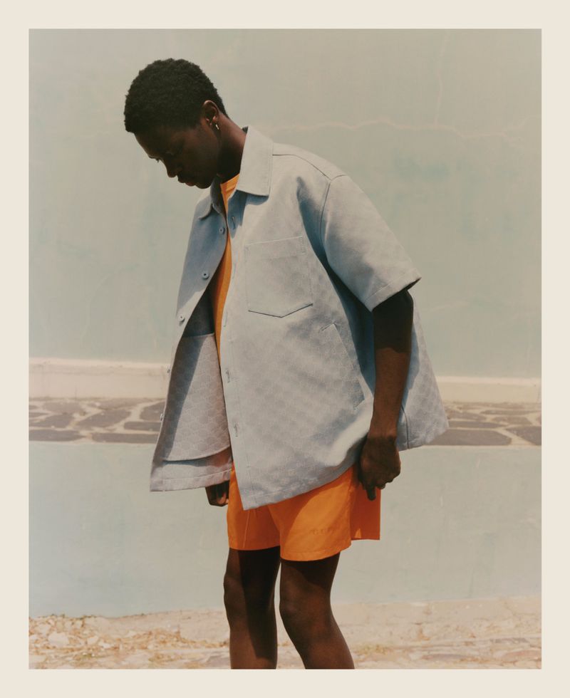 Dark-skinned adult male wearing a light blue button-down shirt and bright orange shorts in an outdoor setting with muted colors.