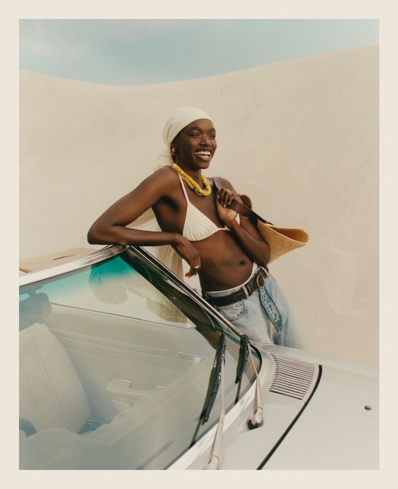 Smiling Black woman in bikini and headscarf leaning against car on sand dune. Car is cream-colored, with an older vintage aesthetic.
